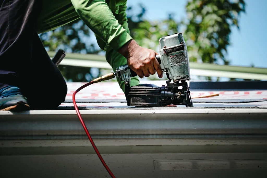 Worker using a pneumatic nailer to fasten red and white roof underlayment on a roof deck