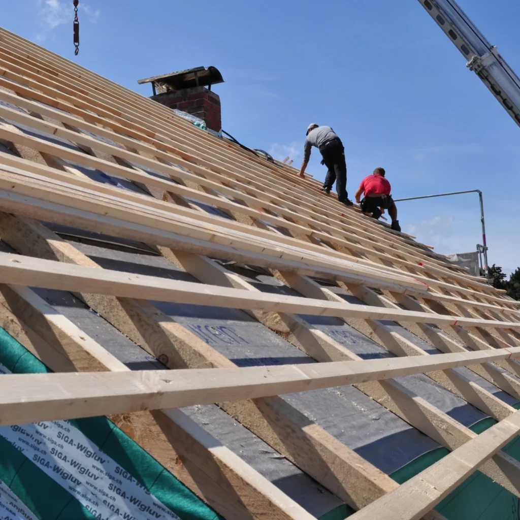 Roof rafters and battens being installed over black synthetic underlayment with a brick chimney and workers on the roof