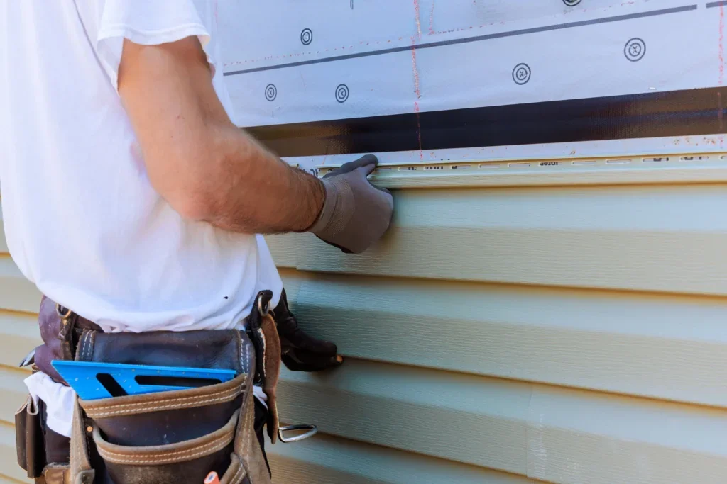 Worker installing pale green vinyl siding over white house wrap with blue speed square in tool belt