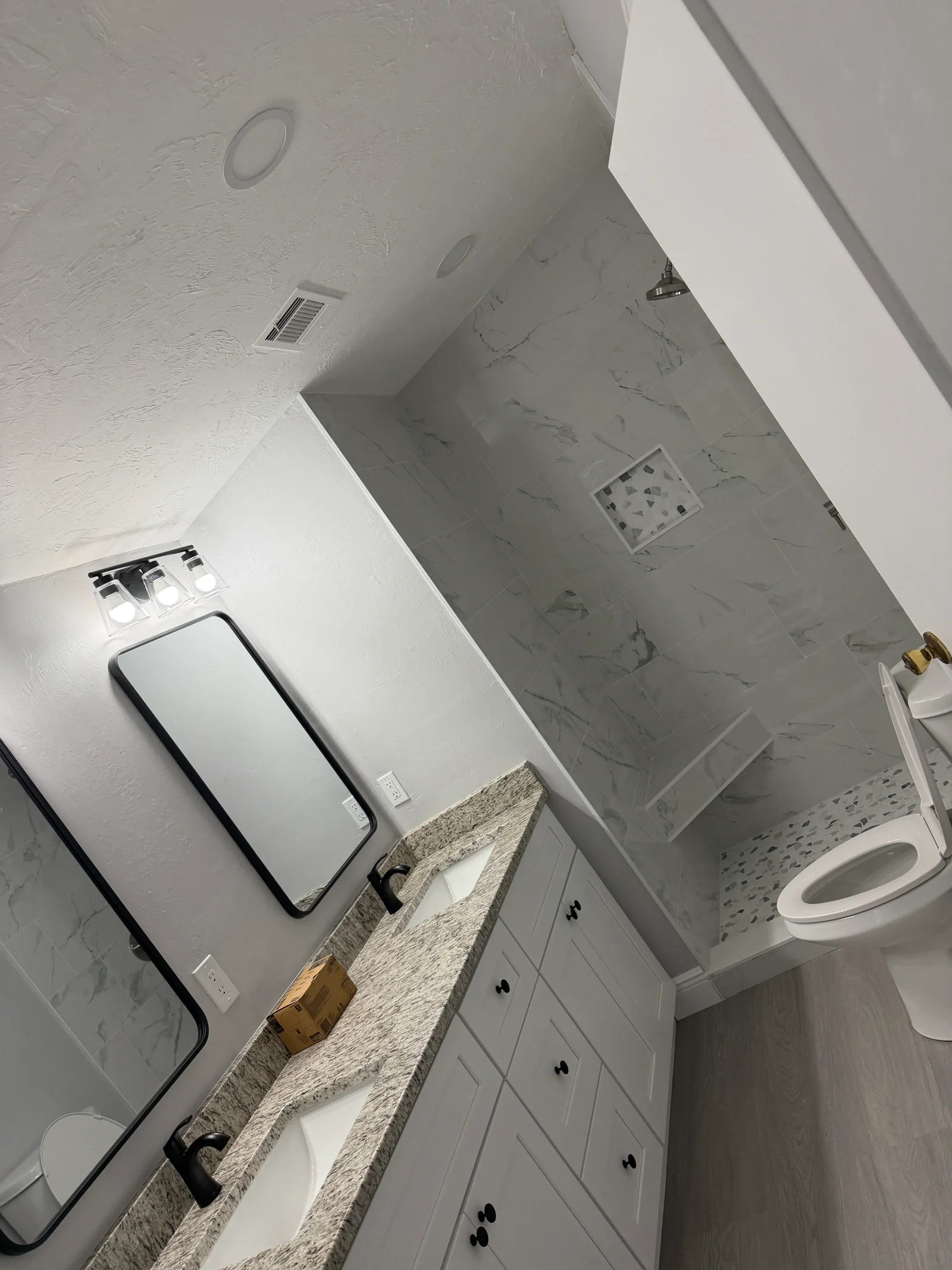 Bathroom with marble-look shower tile, terrazzo niche, granite double vanity, white shaker cabinets, and black framed mirrors