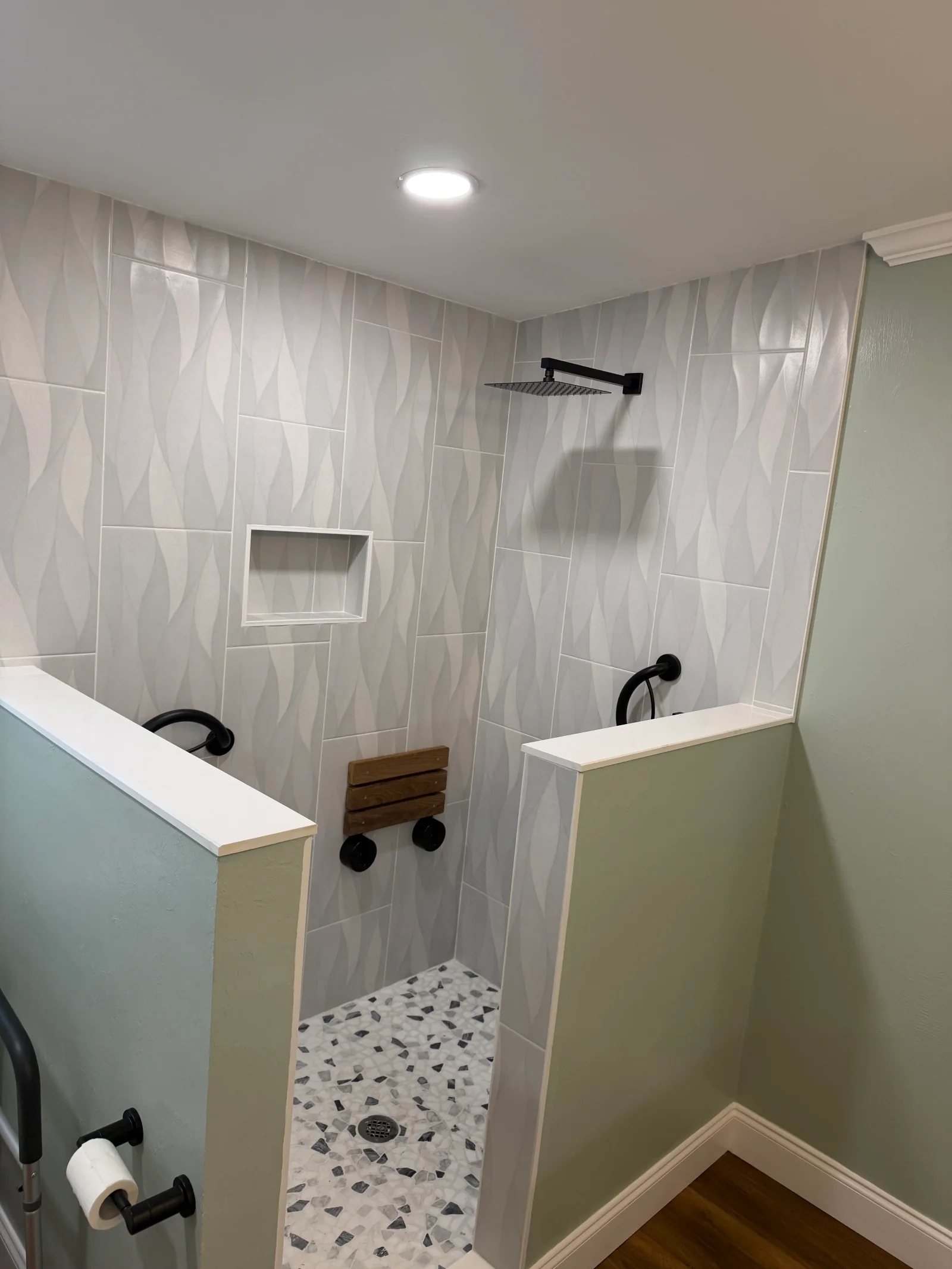 Walk-in shower with wave-textured gray wall tile, terrazzo-style floor, black fixtures, and fold-down teak bench