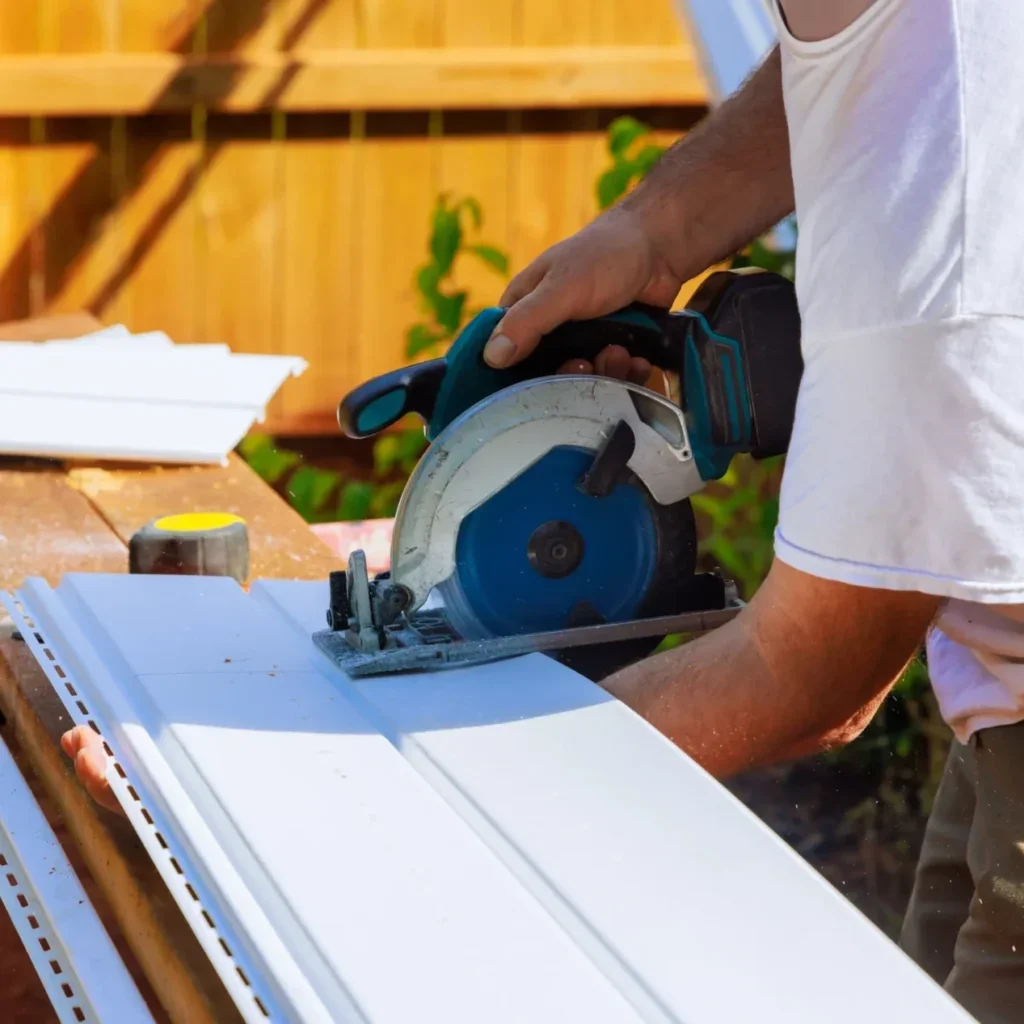 Cordless circular saw cutting white vinyl siding panel on a wooden work surface