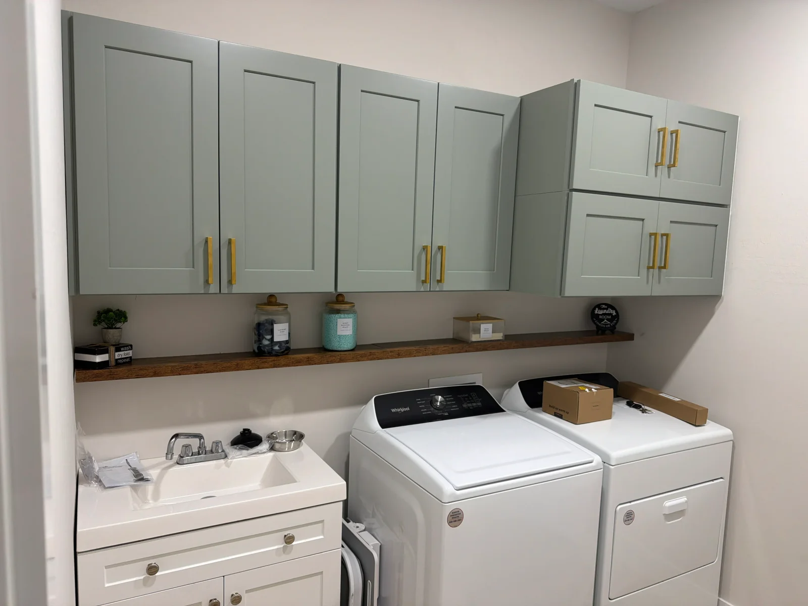 Sage green shaker cabinets with gold pulls above white vanity, floating wood shelf, and white washer and dryer