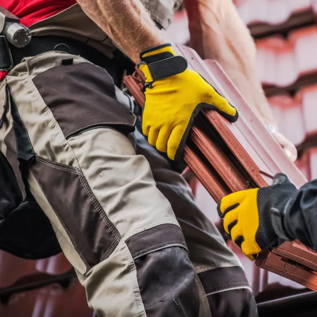 Worker in yellow gloves carrying terracotta clay roof tiles across a steep tiled roof
