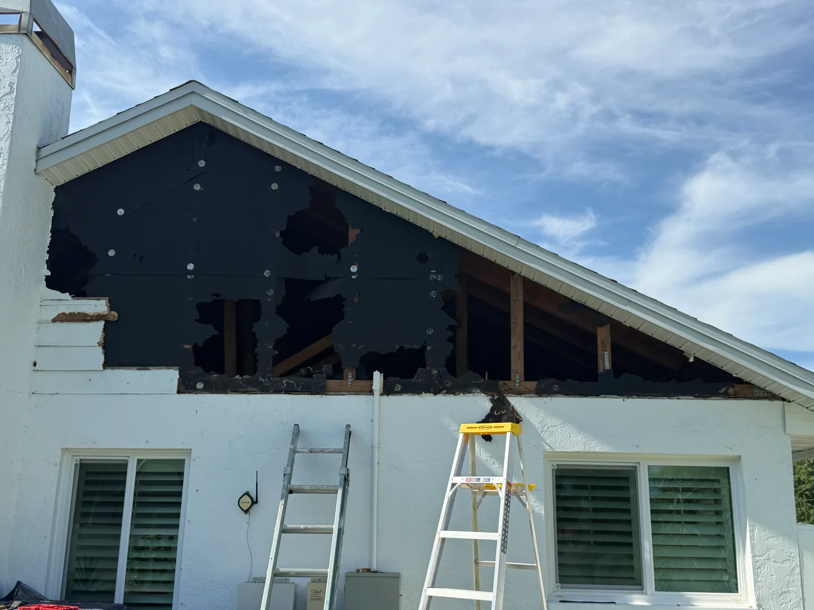 Gable end wall with exposed wood framing, torn black underlayment, and white stucco exterior below, with ladders for access