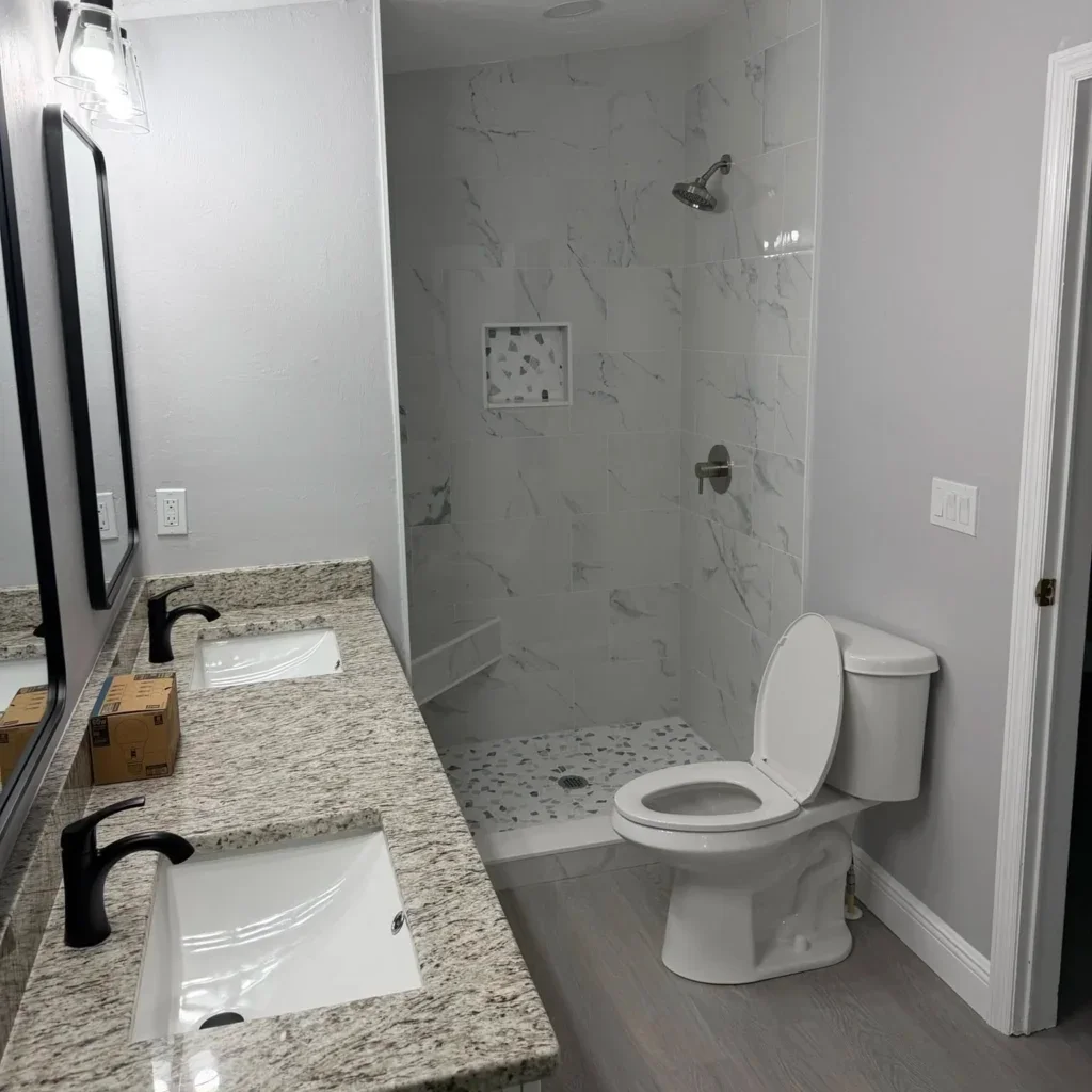 Remodeled bathroom with marble-look shower tile, terrazzo niche, granite double vanity, black faucets, and gray vinyl plank floor