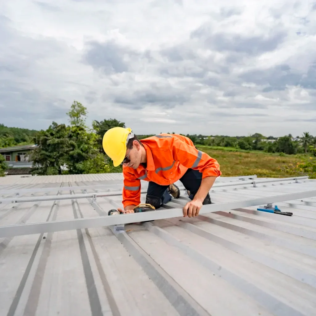Worker in orange hi-vis shirt and hard hat installing silver metal rails on a corrugated metal roof with a cordless drill